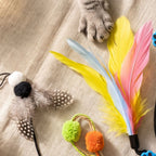 A set of cat toys arranged on a beige fabric surface, including a feather toy with yellow, pink, and light blue feathers, a feather pom-pom toy with black and white spotted feathers, and a small toy with green and orange pom-poms.